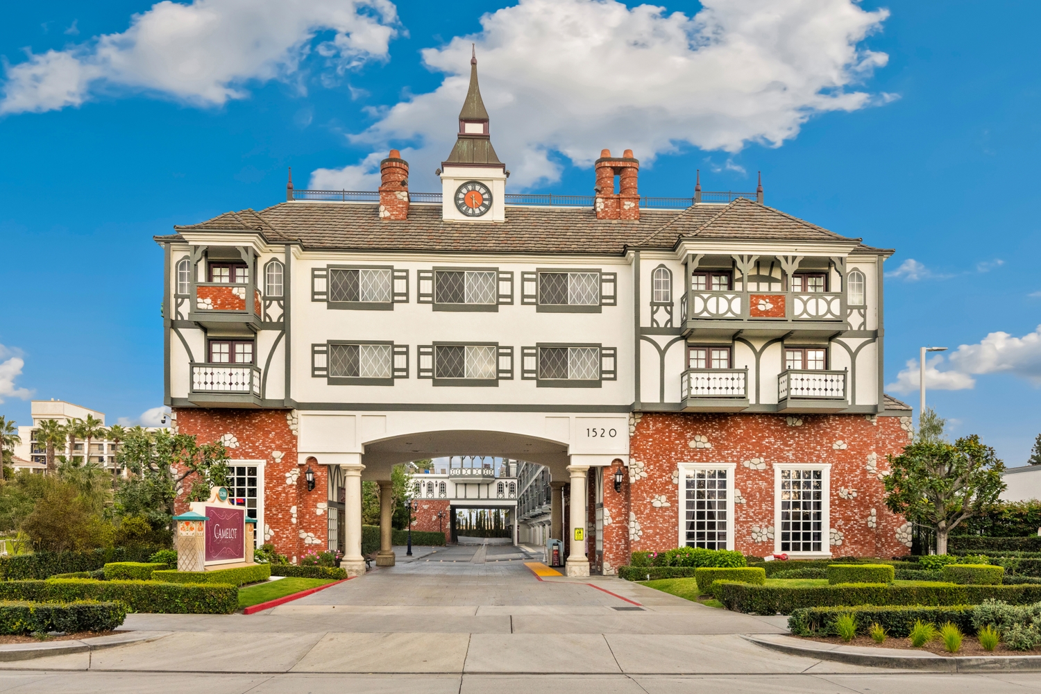 A large, multi-story hotel building with a clock tower, archway, and gardens, set against a blue sky with clouds.